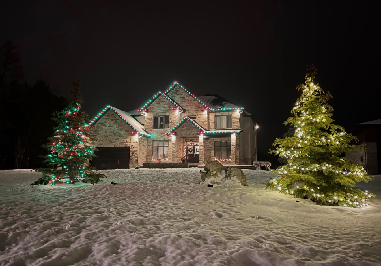 Home with red and green holiday roofline lights beside two lit evergreen trees in the snow (stock image)